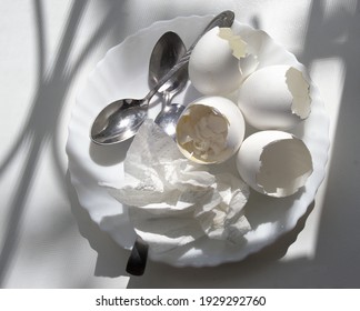 Shells Of Boiled Eggs With The Remains Of Yolk And Boiled Egg White, Used Paper Napkin, Teaspoons, All On White Plate, Under The Sun From Window On A Clean Plastic Windowsill With Picturesque Shadows