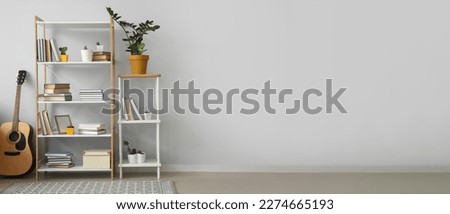 Shelving unit with books, houseplants and guitar near light wall in room. Banner for design
