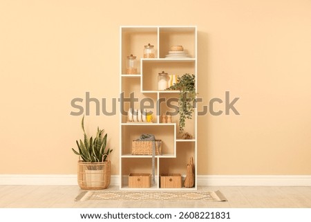 Shelving unit with dishware, jars of different products and other stuff near beige wall indoors