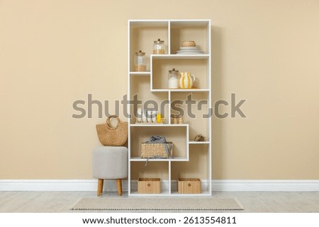 Shelving unit with dishware, jars of different products and other stuff near beige wall indoors