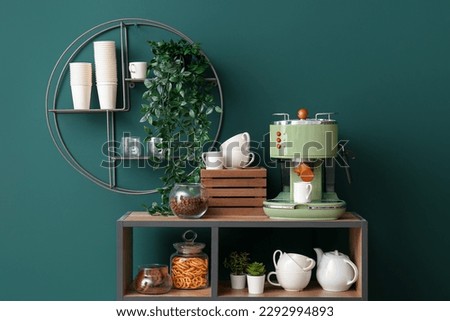 Shelving unit with modern coffee machine, cups and snacks near green wall