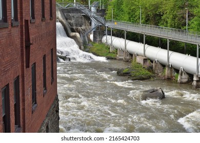 SHERBROOKE, QUEBEC, CANADA - MAY 6, 2022 - Magog River Sherbrooke Abenakis Hydroelectric Power Plant Dam. Large White Penstock And Footbridge