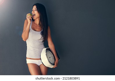 She's All About Healthy Eating. Shot Of A Young Woman Eating An Apple While Holding A Scale.