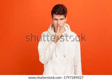 Shh sign. Concerned man holding finger near lips and showing silence sign. Keeping secrets, deceit. Indoor studio shot isolated on orange background