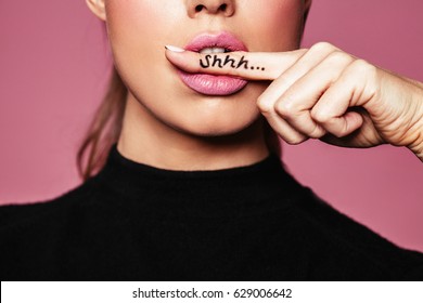 Shh! Women's secrets. Cropped shot of female with finger in mouth. Closeup portrait of young woman is showing a sign of silence with shhh written on the finger.