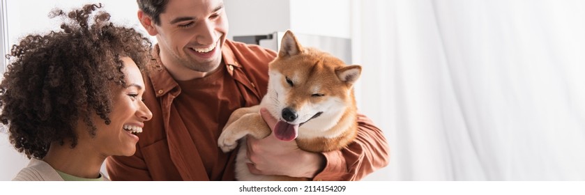 shiba inu dog sticking out tongue near joyful interracial couple in kitchen, banner