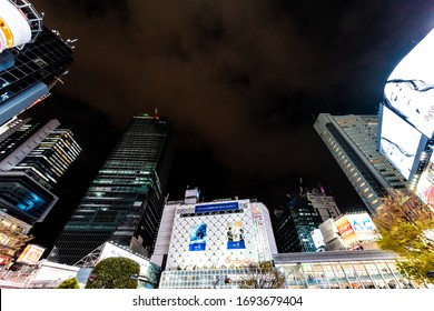 Shibuya, Japan - April 1, 2019: Looking Up Low Angle Wide Angle View Of Tokyo Cityscape At Famous Scrambled Crossing Crosswalk In Downtown City With Neon Bright Lights And Advertisements At Night