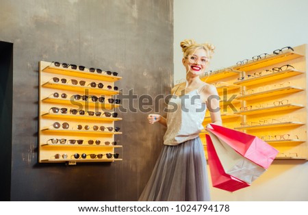 Shining fashion woman with red lips and funny hairstyle holding pink paper bags after bying sunglasses in optical store - Beautiful girl wearing eyeglasses in optician shop. Ophthalmology concept.