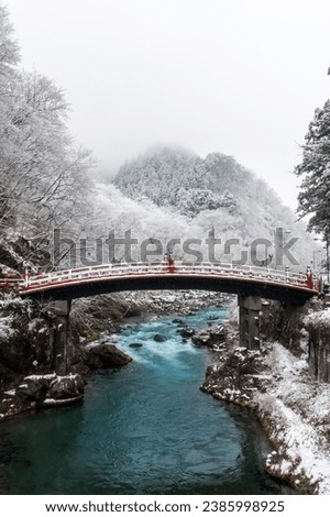 Shinkyo Sacred Bridge red, ancient architecture made from curved shaped wood spanning Daiya River, in winter covered by snow light snowfall. Tourist attraction and landmark in Nikko, Tochigi, Japan.