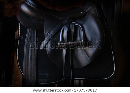 Shiny black leather horse saddle in the dark stable , with stirrup, ready for riding 