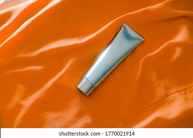 Shiny Light Blue Cosmetic Tube On Sunny Orange Fabric Background. Summer Beauty Product Concept. Blank Space For Text, Brand Packaging, Copy Space. Flatlay