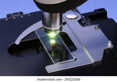 Shiny metal objective and the drop of a liquid illuminated from below by green light on a slide of a dark (obscured) microscope. Focus on full depth. 
