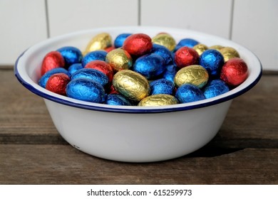 shiny wrapped chocolate Easter eggs, colors red, blue and golden. white enamel bowl filled with decorative Easter eggs. On a wooden background in white and brown colors. festive easter background - Powered by Shutterstock