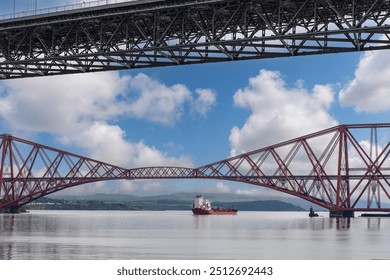 Ship sailing under the red Forth cantilever railway bridge across the Firth of Forth, Queensferry, UK and new Forth Road Bridge crossing in top of image near Edinburgh