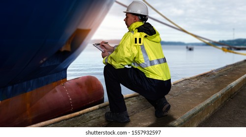 Ship Supervisor Engineer Inspector Stands At The Dockside In A Port. Wearing Safety Helmet And Yellow Vest. Cargo Shipping Industry. Protection And Idemnity Concept.