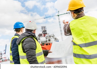 Shipbuilding Engineers Introducing New Solution In A Shipyard. All Men Wearing Safety Helmets And Yellow Vests. Shipbuilding Industry.