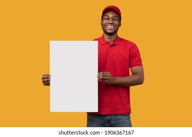 Shipment Service Promotion. Portrait Of Cheerful Bearded Black Delivery Man In Red Uniform Holding And Showing Vertical Empty Blank Board Sign Standing Isolated On Orange Studio Background