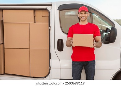 Shipping service concept. Young happy deliveryman with parcel standing near car full of cardboard boxes, smiling at camera - Powered by Shutterstock