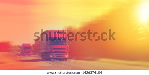 shipping two\
red cargo trucks on the road being driven rays  sun sunset yellow\
evening Cargo delivery. Highway\
road.