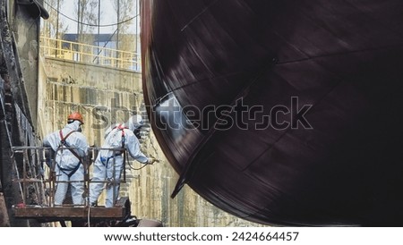 Shipyard Painters apply paint to keel of a large ship while standing on cherry picker working at heights