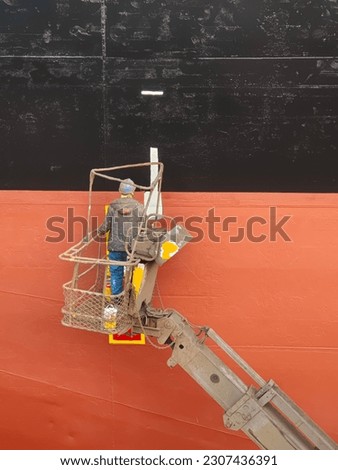 A shipyard worker on a cherry picker is painting on a ship side