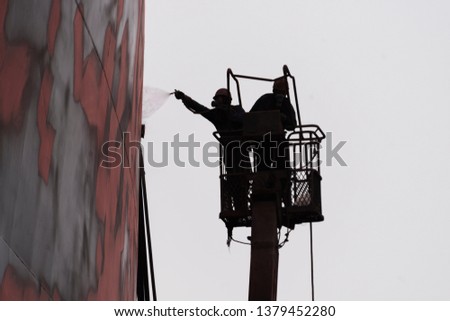 Shipyard workers applying paint coat on a ship's hull for protection against corrosion on a cherry picker by using a spray gun.