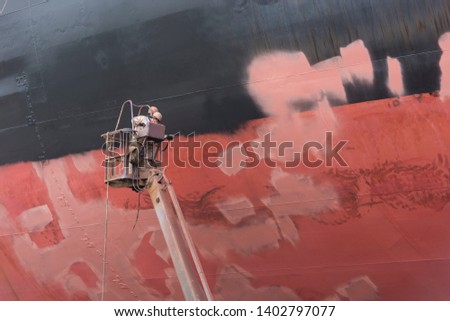 Shipyard workers applying paint coat on a ship's hull for protection against corrosion on a cherry picker by using a spray gun.