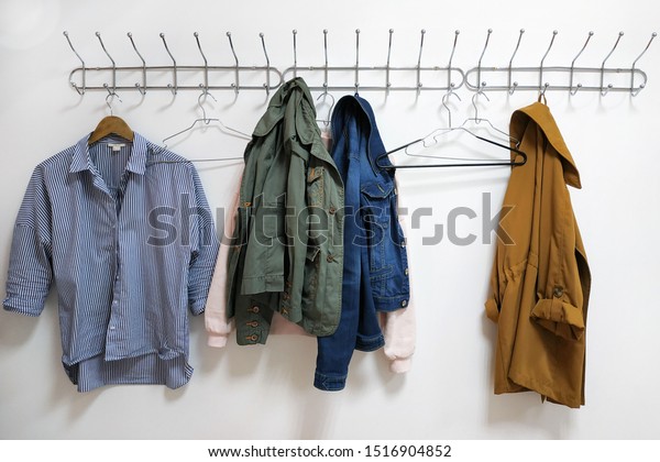 A shirt, jackets and a hanger hang on a metal\
wall hanger. Women\'s clothes hanging on a wall hanger. Hooks with\
clothes hangers on a white\
wall.