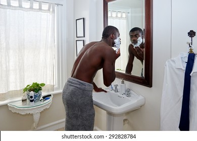 Shirtless African Black Man Applying Shaving Cream To Face In Mirror Reflection For Morning Clean Shaven Look In Home Bathroom