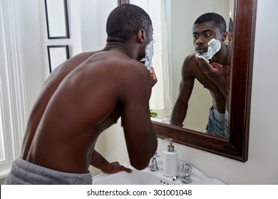 Shirtless African Black Man Applying Shaving Cream To Face In Mirror Reflection For Morning Clean Shaven Look In Home Bathroom