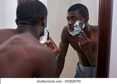 Shirtless African Black Man Shaving Beard Stubble Face With Razor In Mirror Reflection For Morning Clean Shaven Look In Home Bathroom