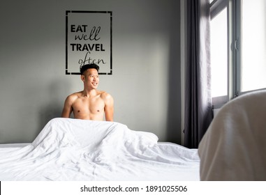 Shirtless Asian Man Sitting In Bed Looking Out The Window Where The Light Comes In, Behind Him There Is A Black Frame Painted On The Wall With The Writing Eat Well Travel Often.