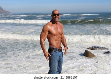 Shirtless Bald Muscle Man In Jeans Showing Off Body Against Ocean Backdrop