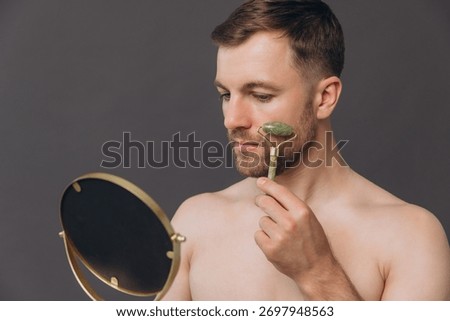 Shirtless bearded man using jade roller during facial massage looking into mirror isolated on gray background, copy space