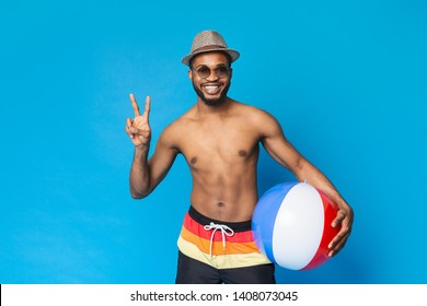Shirtless black guy holding inflatable ball, showing v sign and smiling at camera, blue studio background