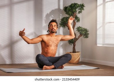 Shirtless Black Man With Closed Eyes Gesticulating While Sitting In Padmasana Pose Near Potted Tree And Meditating During Yoga Session In Morning At Home