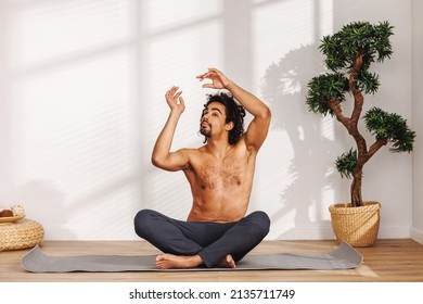 Shirtless Black Man With Closed Eyes Gesticulating While Sitting In Padmasana Pose Near Potted Tree And Meditating During Yoga Session In Morning At Home