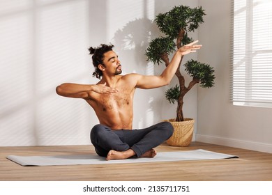 Shirtless Black Man With Closed Eyes Gesticulating While Sitting In Padmasana Pose Near Potted Tree And Meditating During Yoga Session In Morning At Home