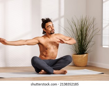 Shirtless Black Man With Closed Eyes Gesticulating While Sitting In Padmasana Pose Near Potted Tree And Meditating During Yoga Session In Morning At Home