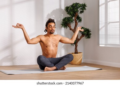 Shirtless Black Man With Closed Eyes Gesticulating While Sitting In Padmasana Pose Near Potted Tree And Meditating During Yoga Session In Morning At Home