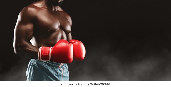 A shirtless boxer wearing red boxing gloves stands in a fighting stance against a black background with smoke. - Powered by Shutterstock