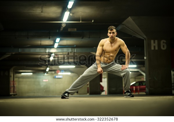 Shirtless handsome muscular young man stretching\
at parking garage and looking at camera, natural lights, dark\
place. Shallow depth of\
field.