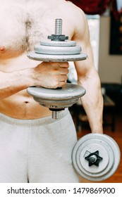 Shirtless Man In Grey Sweat Pants Working Out At Home. Holding Weights In His Arms And Flexing Muscles.