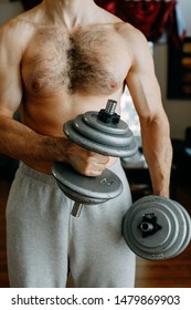 Shirtless Man In Grey Sweat Pants Working Out At Home. Holding Weights In His Arms And Flexing Muscles.