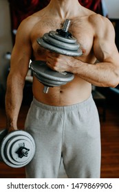 Shirtless Man In Grey Sweat Pants Working Out At Home. Holding Weights In His Arms And Flexing Muscles.