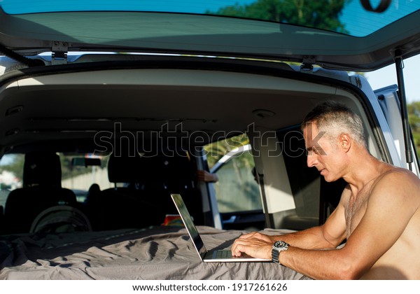 Shirtless man in holiday setting works on the\
computer in the hood of his\
van