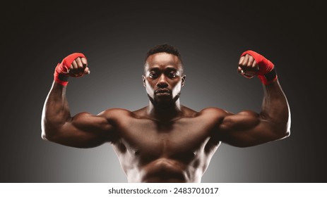 A shirtless man with red hand wraps flexes his bicep muscles in a studio setting. - Powered by Shutterstock