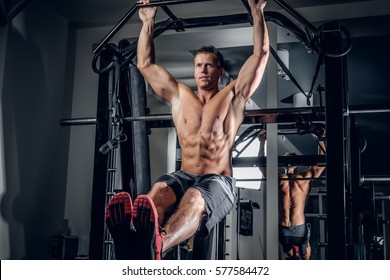 Shirtless Muscular Man Doing ABS Workouts On Pull Up Bar In A Gym.