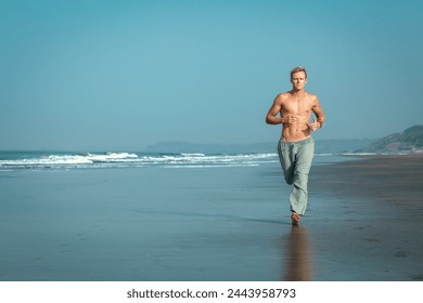 A shirtless, muscular man jogs along the wet sand of a beach, with the ocean and mountains in the background. He has an intense, focused expression on his face. Jogging on the sea beach. - Powered by Shutterstock