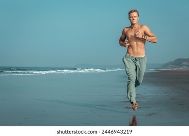 A shirtless, muscular man jogs along the wet sand of a beach, with the ocean and mountains in the background. He has an intense, focused expression on his face. Jogging on the sea beach. - Powered by Shutterstock